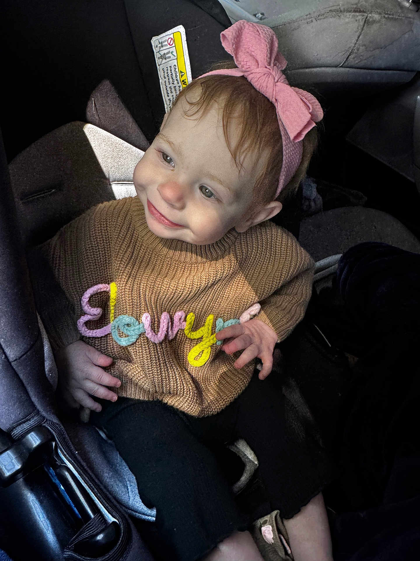 Elowyn is registered to the contest to win money with this photo: baby, brown_sweater, car_seat, child, clothing, colorful_letters, cute, hand, happy, headband, indoor, infant, person, pink_bow, portrait, seatbelt, smiling, sunlight, sweater, toddler