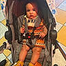 Rmani is registered to the contest to win money with this photo: baby, stroller, child, footwear, sandals, blanket, patterned_clothing, floor_tiles, seatbelt, curious_expression, indoor, baby_clothes, legs, hands, face, person, cute, child_seat, sitting, colorful