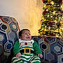 baby, celebration, child, christmas_tree, couch, cozy, decorations, festive, green_outfit, happy, holiday, home, indoor, infant, lights, patterned_fabric, portrait, red_socks, seated, smiling
