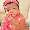 Valerie is registered to the contest to win money with this photo: baby, infant, pink_clothing, heart_pattern, headband, hand, ring, person, face, portrait, newborn, cute, expression, skin, fingernails, holding, soft_background, indoors, closeup, child