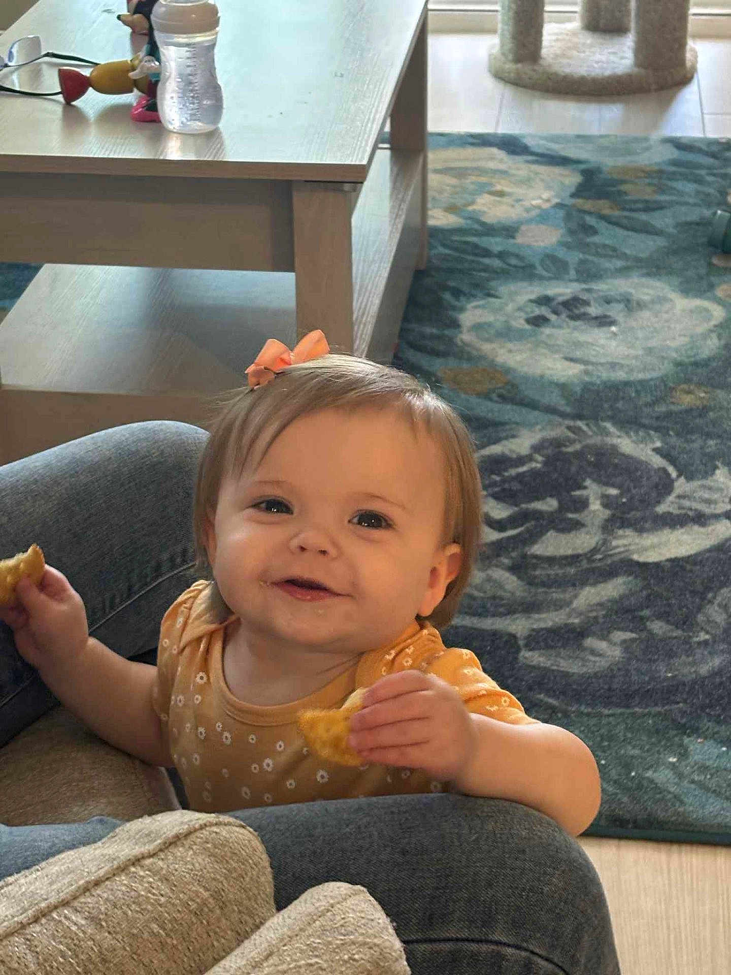 Annie is registered to the contest to win money with this photo: toddler, child, smiling, eating, snack, bow, hair_accessory, clothing, jeans, furniture, table, carpet, rug, floor, indoor, person, happy, cute, cozy, home