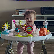 Annie is registered to the contest to win money with this photo: baby, child, activity_walker, toy, indoor, floor, pink_onesie, books, television, smiling, happy, person, furniture, carpet, legs, face, hair, play, home, living_room
