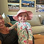 Annie is registered to the contest to win money with this photo: baby, child, woman, smile, hat, flamingo, clothing, couch, indoor, painting, wall_art, living_room, person, happy, portrait, casual, furniture, home, relaxation, sun_hat
