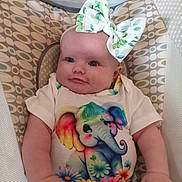 Thora is registered to the contest to win money with this photo: baby, child, infant, bow, onesie, elephant, colorful, patterned, seat, cushion, smiling, cute, happy, portrait, indoors, clothing, headwear, flower, skin, person