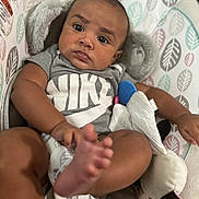 Adonis is registered to the contest to win money with this photo: baby, infant, child, nike, grey_shirt, foot, toy, plush_toy, baby_seat, patterned_fabric, indoor, closeup, person, cute, young_child, relaxed, expression, brown_skin, short_hair, comfortable