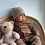 baby, child, teddy_bear, beanie, corduroy, pajamas, wicker_chair, plush_toy, carpet, indoor, cozy, cute, portrait, sitting, soft_toy, warm_clothing, infant, floor, neutral_background, expression