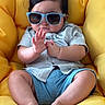 Mahir is registered to the contest to win money with this photo: baby, child, sunglasses, blue_shorts, shirt, yellow_chair, cushion, indoors, cute, chubby, hand_gesture, relaxed, fashion, portrait, person, skin, feet, hair, casual, comfortable