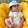 baby, infant, child, yellow_chair, cushion, hat, romper, fruit_print, sitting, cute, indoor, portrait, skin, face, hand, foot, clothing, curious, young, comfortable