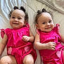 baby, twins, smiling, pink_clothing, heart_patch, curly_hair, hair_bun, earrings, lying_down, white_quilt, infant, cute, happy, portrait, child, indoor, soft_lighting, two_children, matching_outfits, skin