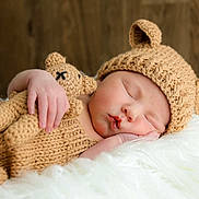 Bentley joined the competition — help win amazing prizes! baby, sleeping, knitted_hat, knitted_outfit, teddy_bear, knitted_toy, cuddling, cozy, cute, infant, child, soft_texture, fluffy, white_blanket, wooden_background, portrait, peaceful, resting, closeup, indoors