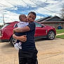 child, baby, outdoor, holding, car, red_car, driveway, house, sky, clouds, casual_clothing, shorts, tshirt, footwear, family, affection, portrait, person, sibling, love