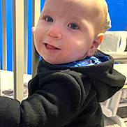 Waylon is registered to the contest to win money with this photo: baby, black_clothing, blue_wall, child, crib, cute, expression, face, furniture, hand, happy, head, home, hoodie, indoor, person, portrait, smile, warm_clothing, young_child