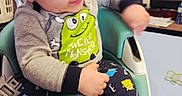 Mateo is registered to the contest to win money with this photo: baby, brown_hair, cartoon, child, clothing, face, furniture, happy, high_chair, indoor, letters, living_room, monster, pajamas, portrait, shirt, sitting, smiling, television, toddler