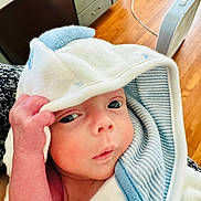 Kase is registered to the contest to win money with this photo: baby, infant, hooded_towel, hand, face, eyes, skin, indoor, wooden_floor, furniture, curious, child, person, cute, newborn, wrapped, soft_texture, striped_towel, closeup, portrait