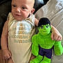 baby, child, plush_toy, hulk, green, onesie, clothing, couch, pillow, infant, cute, smiling, face, person, indoor, soft_toy, relaxing, lying_down, expression, furniture