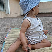 Mensïa participe au concours pour gagner de l'argent avec cette photo : toddler, child, sand, hat, romper, sitting, outdoor, beach_mat, barefoot, hand, legs, wood, curious, summer, casual_clothing, side_view, sun_hat, nature, play, daylight