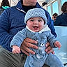 baby, adult, blue_onesie, knitted_hat, smiling, happy, indoor, person, holding, seated, glasses, fleece_jacket, hand, window, daylight, people, transport, casual_clothing, face, portrait