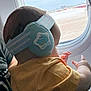 baby, child, earmuffs, airplane_window, yellow_clothing, hand, adult, travel, infant, seat, window, sky, aircraft, head, person, cabin, cute, holding, blue, dinosaur
