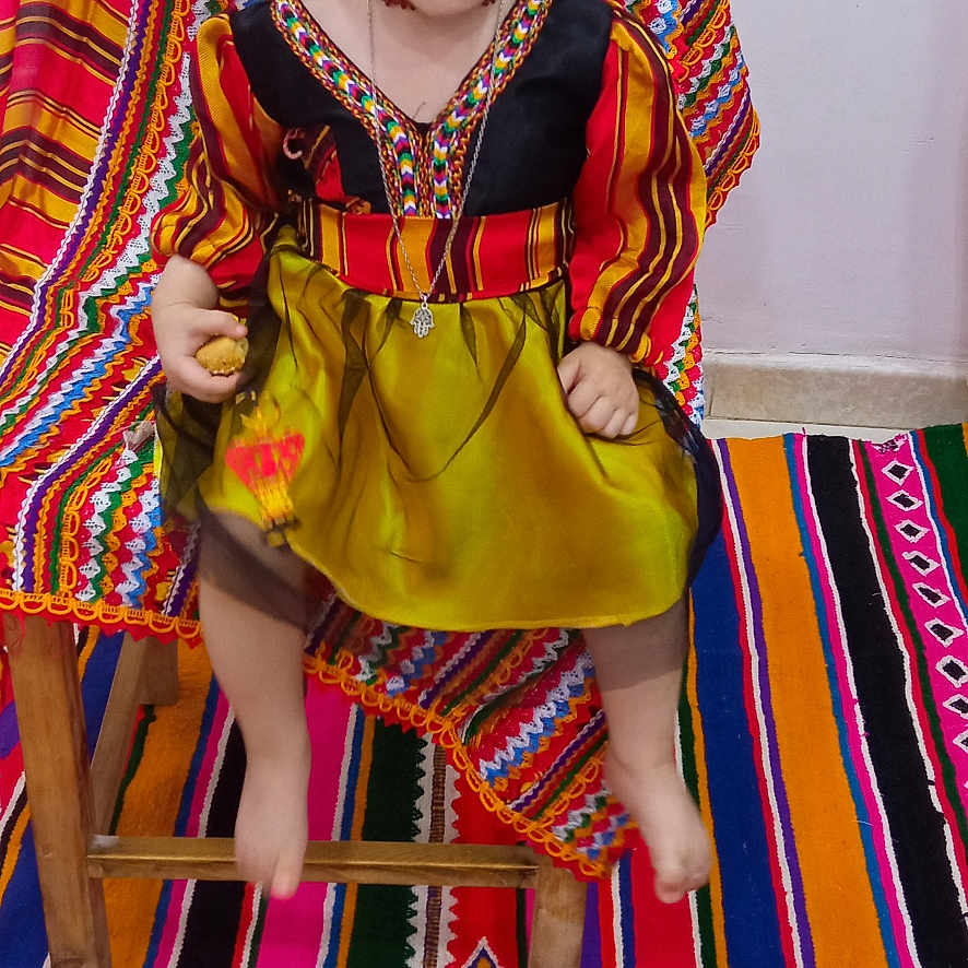 Alissya joined the competition — help win amazing prizes! child, toddler, girl, portrait, sitting, traditional_clothing, colorful_dress, braided_hair, hair_bows, smiling, indoors, chair, woven_textiles, colorful_background, striped_rug, matching_shoes, cultural_attire, full_body, wooden_furniture, photography