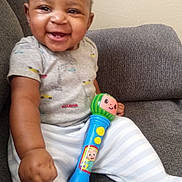 Elijah joined the competition — help win amazing prizes! baby, smiling, toy, microphone, couch, infant, happy, child, clothing, striped_pants, gray_couch, indoors, cute, sitting, person, playful, young_child, casual, short_hair, bright