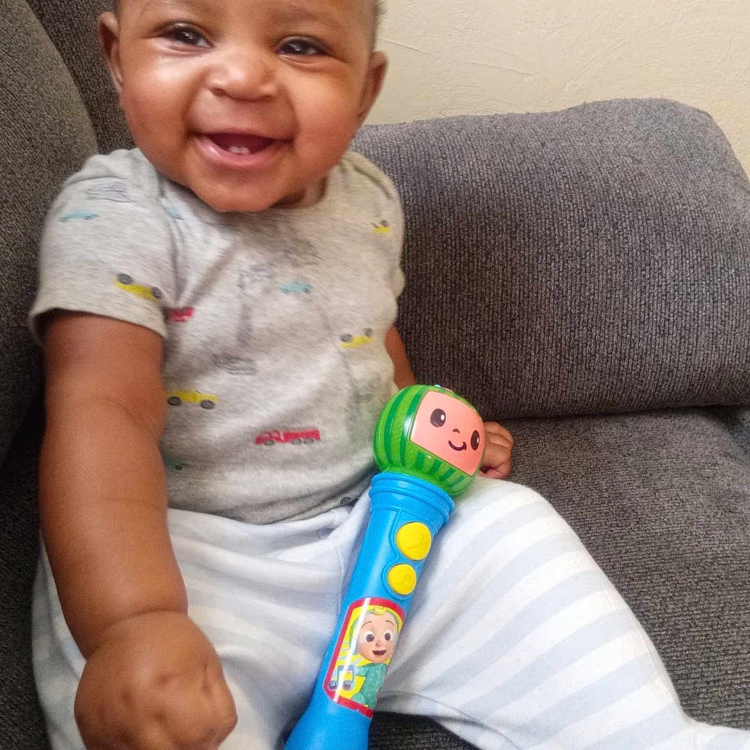 Elijah joined the competition — help win amazing prizes! baby, smiling, toy, microphone, couch, infant, happy, child, clothing, striped_pants, gray_couch, indoors, cute, sitting, person, playful, young_child, casual, short_hair, bright