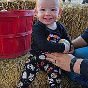 Sophia is registered to the contest to win money with this photo: autumn, black_shirt, casual, child, daylight, festival, fun, halloween_clothing, hay_bale, holding, leggings, outdoor, people, pumpkin, red_bucket, smiling, socks, supporting_hands, toddler, tree