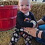 Sophia is registered to the contest to win money with this photo: autumn, black_shirt, casual, child, daylight, festival, fun, halloween_clothing, hay_bale, holding, leggings, outdoor, people, pumpkin, red_bucket, smiling, socks, supporting_hands, toddler, tree