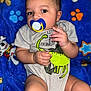 baby, blue_blanket, cartoon_characters, child, close_up, clothing, colorful, cute, dinosaur, expression, face, hand, indoor, infant, legs, lying_down, onesie, pacifier, portrait, soft_texture