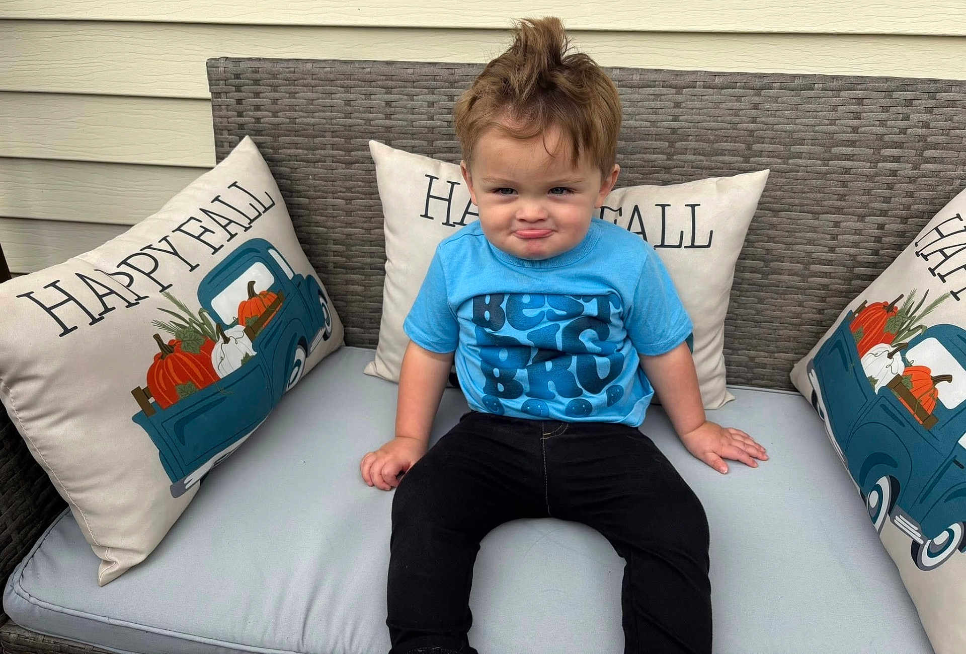Nicolai is registered to the contest to win money with this photo: toddler, child, boy, blue_shirt, black_pants, sitting, outdoor_furniture, cushion, pillow, happy_fall, pumpkin, fall_decor, funny_face, hair_tuft, portrait, cute, smirk, casual_clothing, bench, daylight