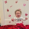 baby, curly_hair, smiling, valentines_day, heart, red_blanket, white_shirt, indoor, child, cute, happy, celebration, holiday, decorations, stickers, infant, portrait, sitting, joyful, festive