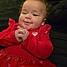 baby, child, red_dress, heart_pattern, smiling, curly_hair, cute, infant, portrait, hands_clasped, happy, indoors, closeup, face, person, toddler, fashion, clothing, sitting, black_background