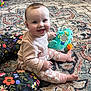 Charlotte is registered to the contest to win money with this photo: baby, infant, child, smiling, sitting, toy, plush_toy, patterned_rug, floral_pillow, checkered_pants, onesie, carpet, living_room, cabinet, playtime, cute, happy_face, cheeks, bare_foot, portrait