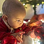 baby, child, christmas_ornaments, red_clothing, holiday_lights, decorations, festive, closeup, face, hands, curious, indoor, soft_light, blurred_background, celebration, winter, warm, cozy, infant, portrait