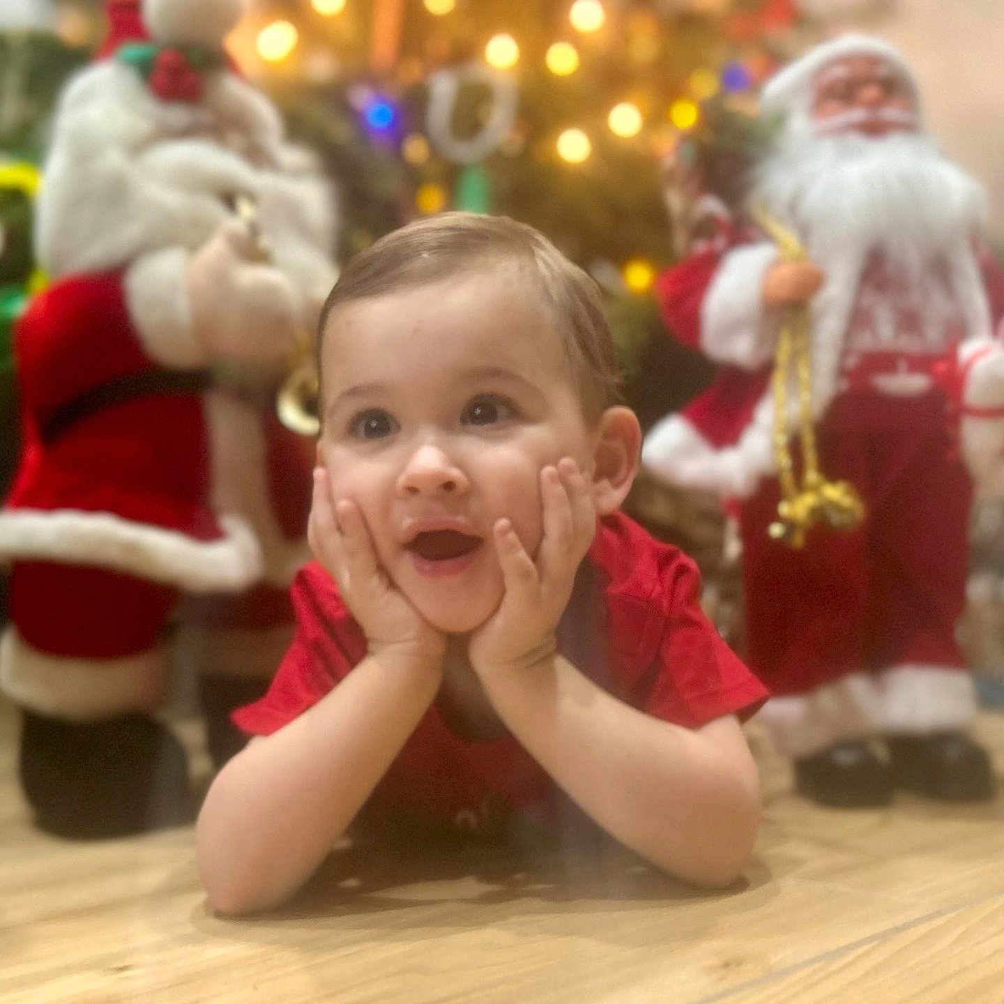 Yliann a rejoint le concours — aidez-le/la à gagner de superbes lots ! blurred_background, celebration, child, christmas, cute, decorations, festive, floor, hands_on_cheeks, happy, holiday, indoors, lights, portrait, red_shirt, santa_claus, seasonal, smiling, toddler, wooden_floor