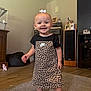 toddler, child, smile, leopard_print, dress, bow, nike_logo, barefoot, standing, tooth, indoor, living_room, carpet, hardwood_floor, speaker, cabinet, picture_frame, toy, happy, portrait