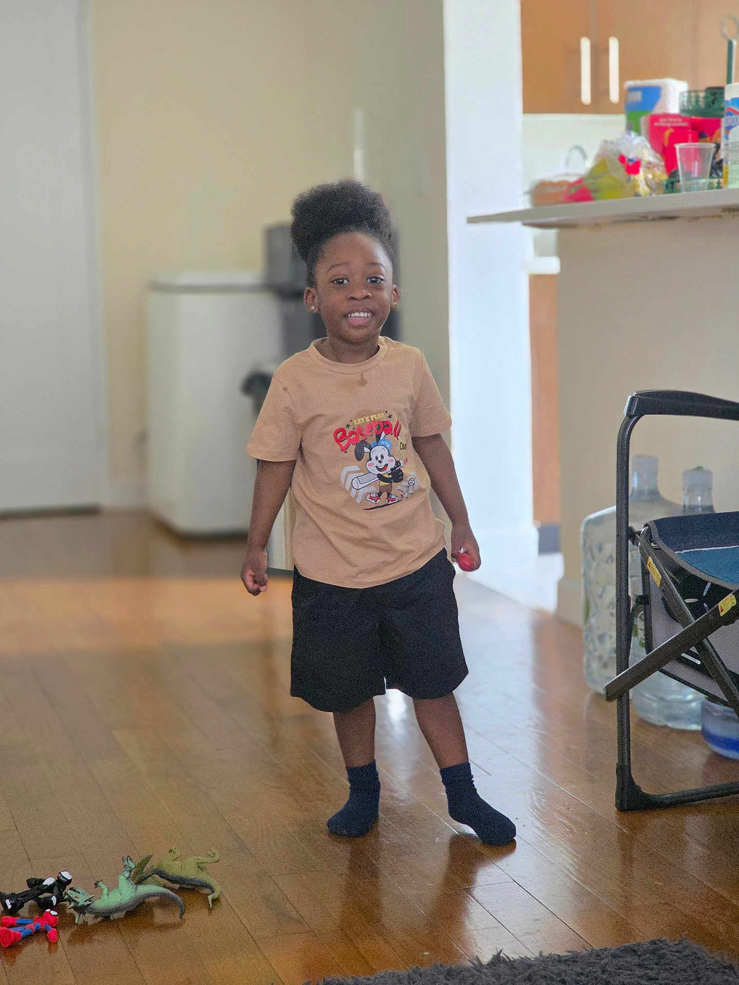Kasen is registered to the contest to win money with this photo: child, indoor, smiling, tshirt, shorts, toy, floor, wooden_floor, living_room, kitchen, furniture, person, happy, playtime, socks, home, young_child, standing, casual_clothing, afro_hair