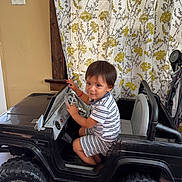 Gael is registered to the contest to win money with this photo: child, toddler, toy_car, indoor, striped_clothing, steering_wheel, smiling, curtains, floral_pattern, floor, wheels, seat, window, person, play, home, fun, vehicle, black, shorts