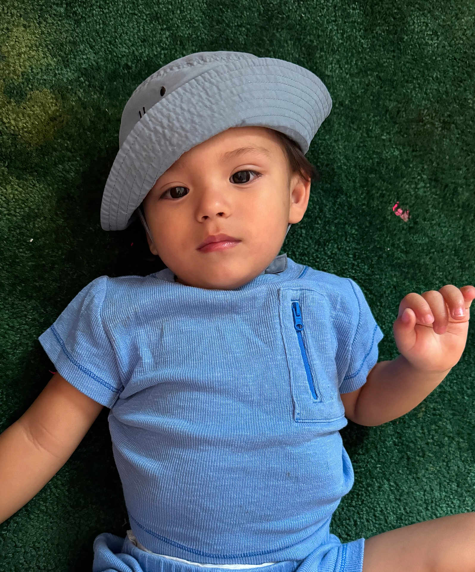 Gael joined the competition — help win amazing prizes! toddler, child, hat, blue_clothing, lying_down, green_background, face, portrait, casual, relaxed, young_child, outdoor_surface, short_sleeves, zipper_pocket, baby, cute, small_hand, expression, headwear, one_person