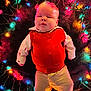 Ace is registered to the contest to win money with this photo: baby, child, infant, christmas_lights, red_vest, white_shirt, pants, barefoot, lying_down, holiday, festive, colorful_lights, cozy, portrait, indoors, cute, smiling, cheeks, warm_lighting, fur_texture