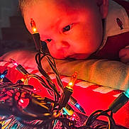 Ace joined the competition — help win amazing prizes! baby, child, face, closeup, christmas_lights, colorful, curious, pillow, red_clothing, indoors, holiday, reflection, string_lights, tangled_wires, soft_lighting, cozy, portrait, young_child, festive, decor