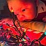 baby, child, face, closeup, christmas_lights, colorful, curious, pillow, red_clothing, indoors, holiday, reflection, string_lights, tangled_wires, soft_lighting, cozy, portrait, young_child, festive, decor