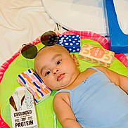 Zainab is registered to the contest to win money with this photo: baby, infant, playmat, sunglasses, protein_drink, carton, colorful_boxes, child, blue_onesie, skin, headwear, indoors, cute, relaxed, lying_down, soft_toys, bright_colors, food_packaging, drink, baby_fashion