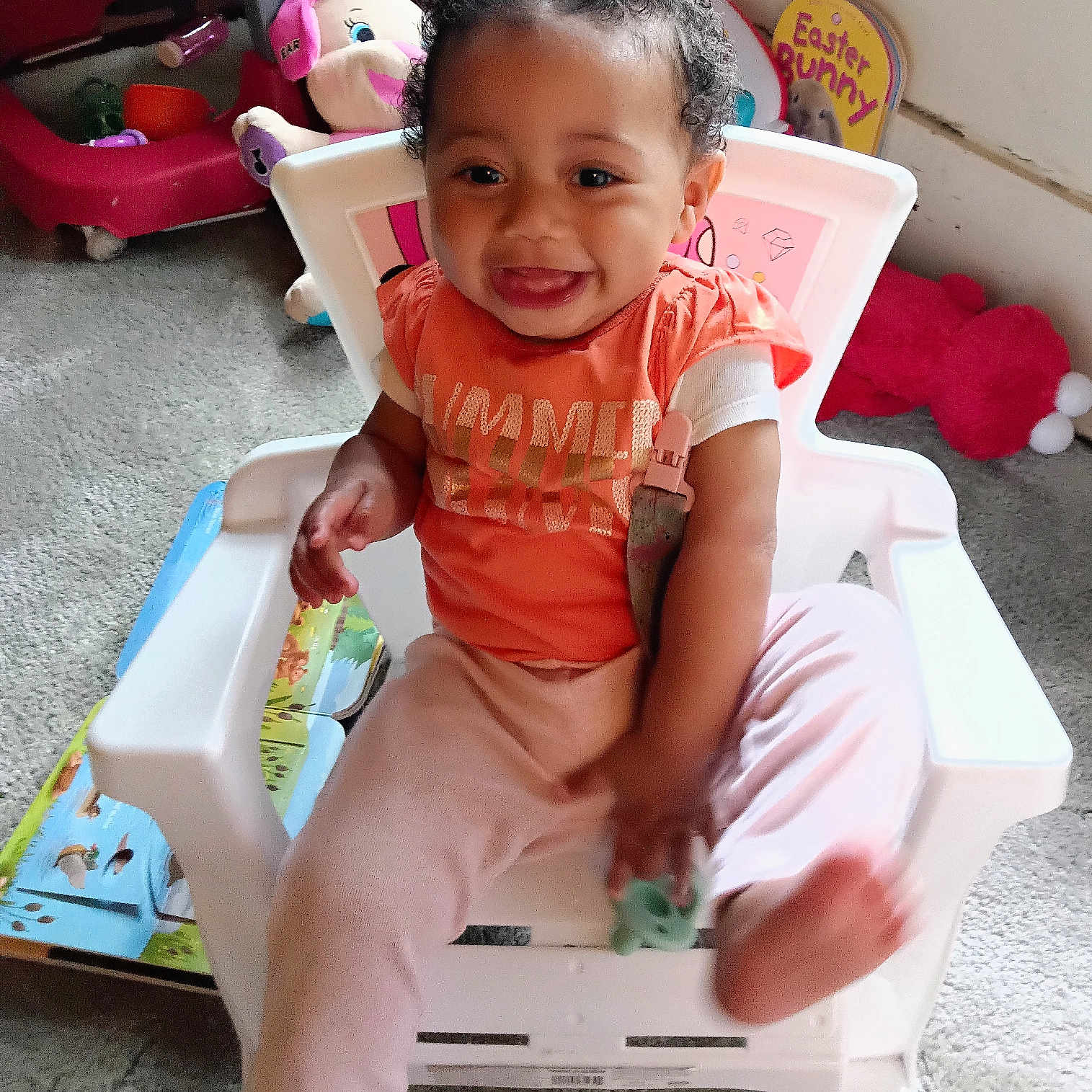 Amina is registered to the contest to win money with this photo: baby, carpet, chair, child, cute, hair_bows, happy, holding_toy, indoor, light_pink_pants, orange_shirt, person, pink_bows, plastic_chair, playroom, sitting, smiling, toddler, toy, young_child