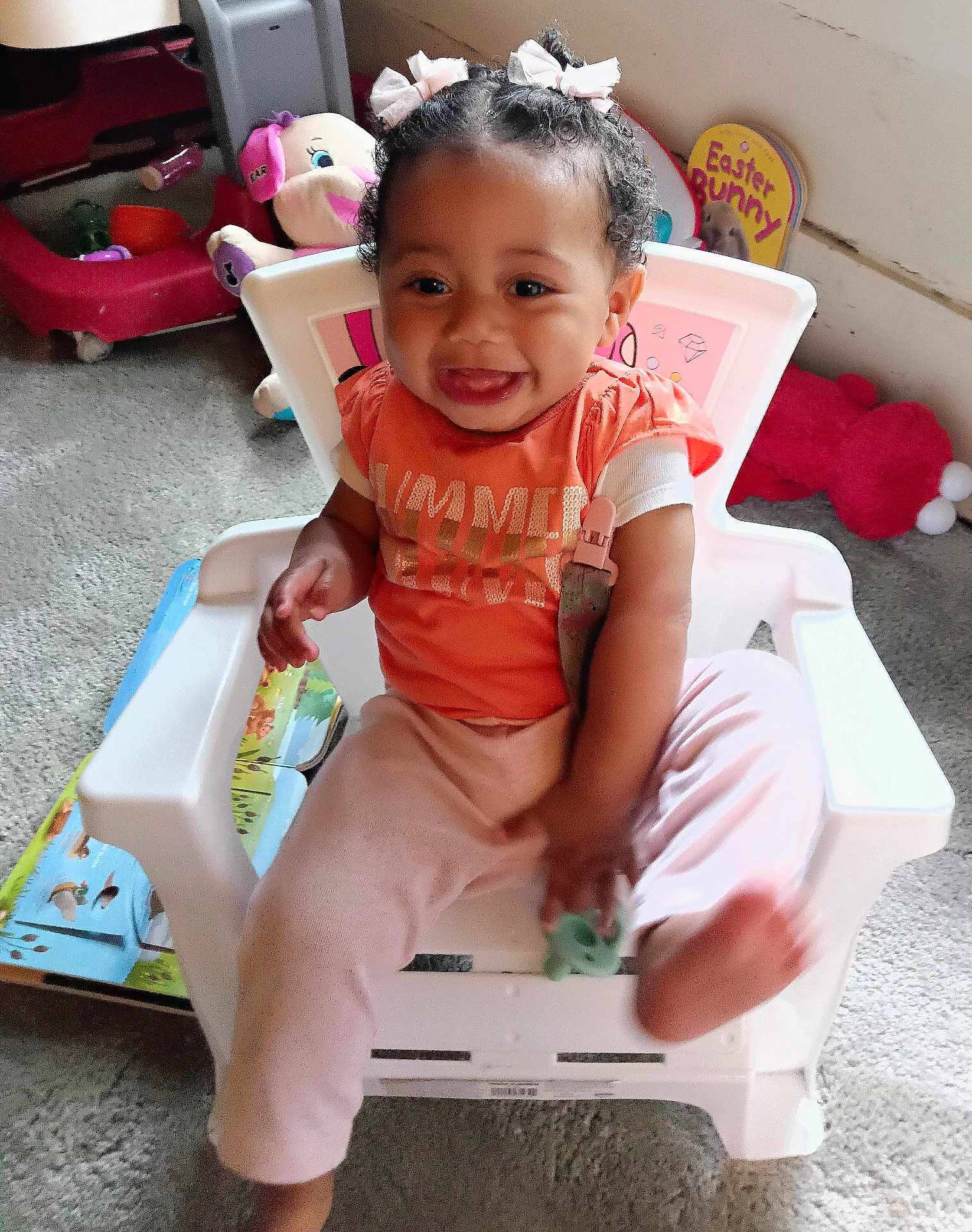 Amina is registered to the contest to win money with this photo: toddler, child, smiling, indoor, chair, toy, pink_bows, orange_shirt, light_pink_pants, happy, cute, hair_bows, carpet, playroom, plastic_chair, baby, person, young_child, sitting, holding_toy
