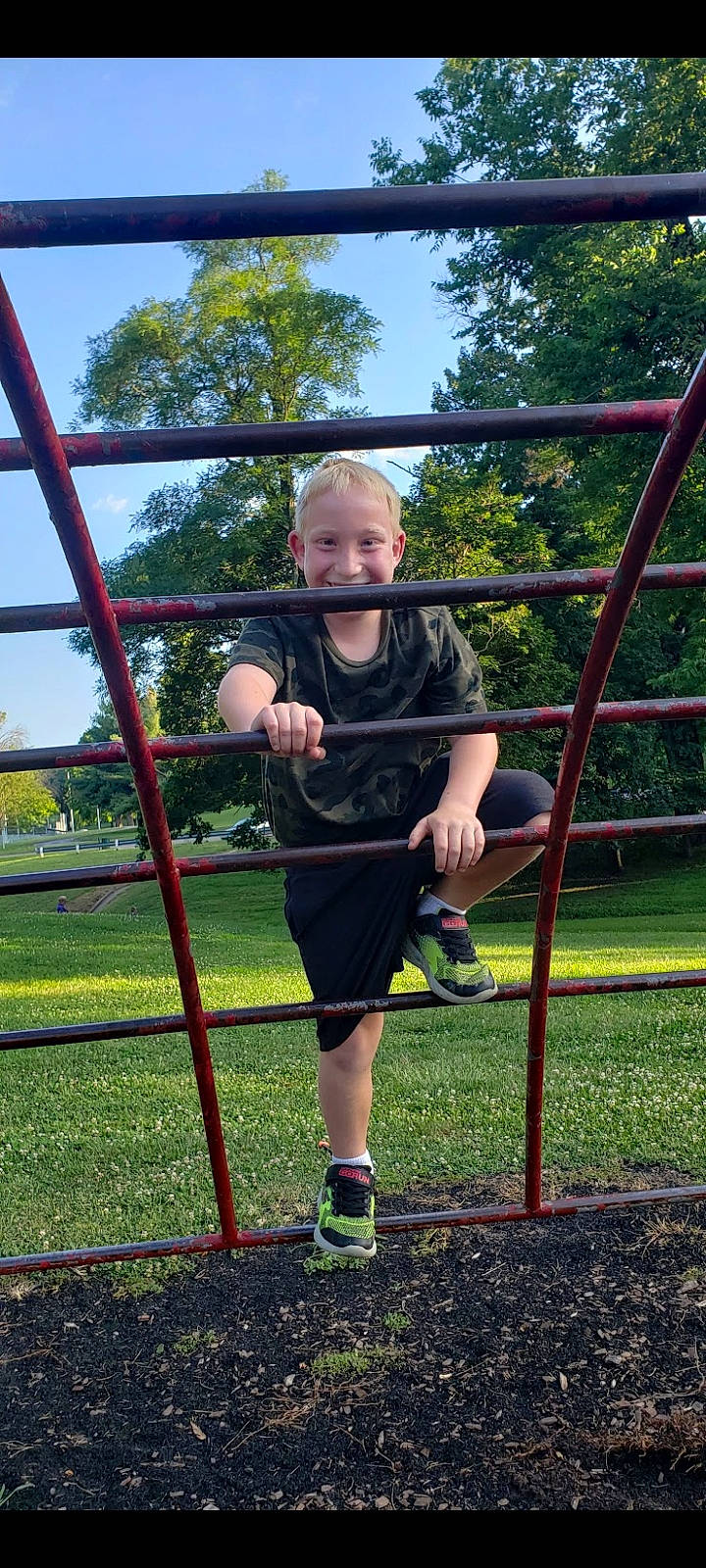 Ethan is registered to the contest to win money with this photo: city, fence, footwear, fun, grass, joy, leg, leisure, outdoor_play_equipment, person, plant, playground, public_space, recreation, shorts, sky, smile, sneakers, swing, t_shirt