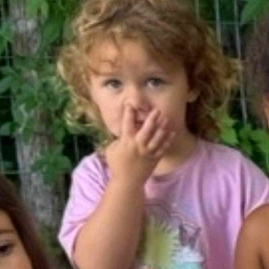 Piper is registered to the contest to win money with this photo: casual_clothing, child, curly_hair, cute, daylight, expression, fence, friends, greenery, kids, nature, nose_picking, outdoor, people, pink_shirt, playful, portrait, sitting, summer, toddler