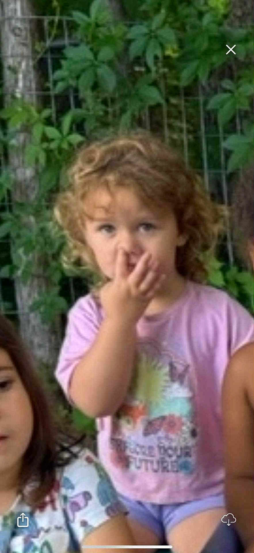 Piper is registered to the contest to win money with this photo: child, toddler, curly_hair, pink_shirt, nose_picking, outdoor, greenery, fence, kids, casual_clothing, portrait, summer, nature, cute, playful, expression, sitting, people, friends, daylight