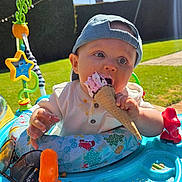 Matteo is registered to the contest to win money with this photo: baby, ice_cream, cap, outdoor, sunlight, baby_walker, toy, grass, hedge, summer, blue_sky, child, cute, eating, cone, playtime, sunshine, smiling, colorful, fun