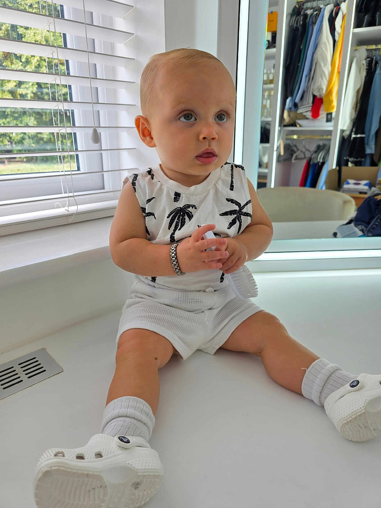 Matteo joined the competition — help win amazing prizes! toddler, child, indoor, window, blinds, white_clothing, shorts, socks, white_shoes, bracelet, holding_object, hairbrush, closet, clothes, sitting, curious, face, skin, light, home