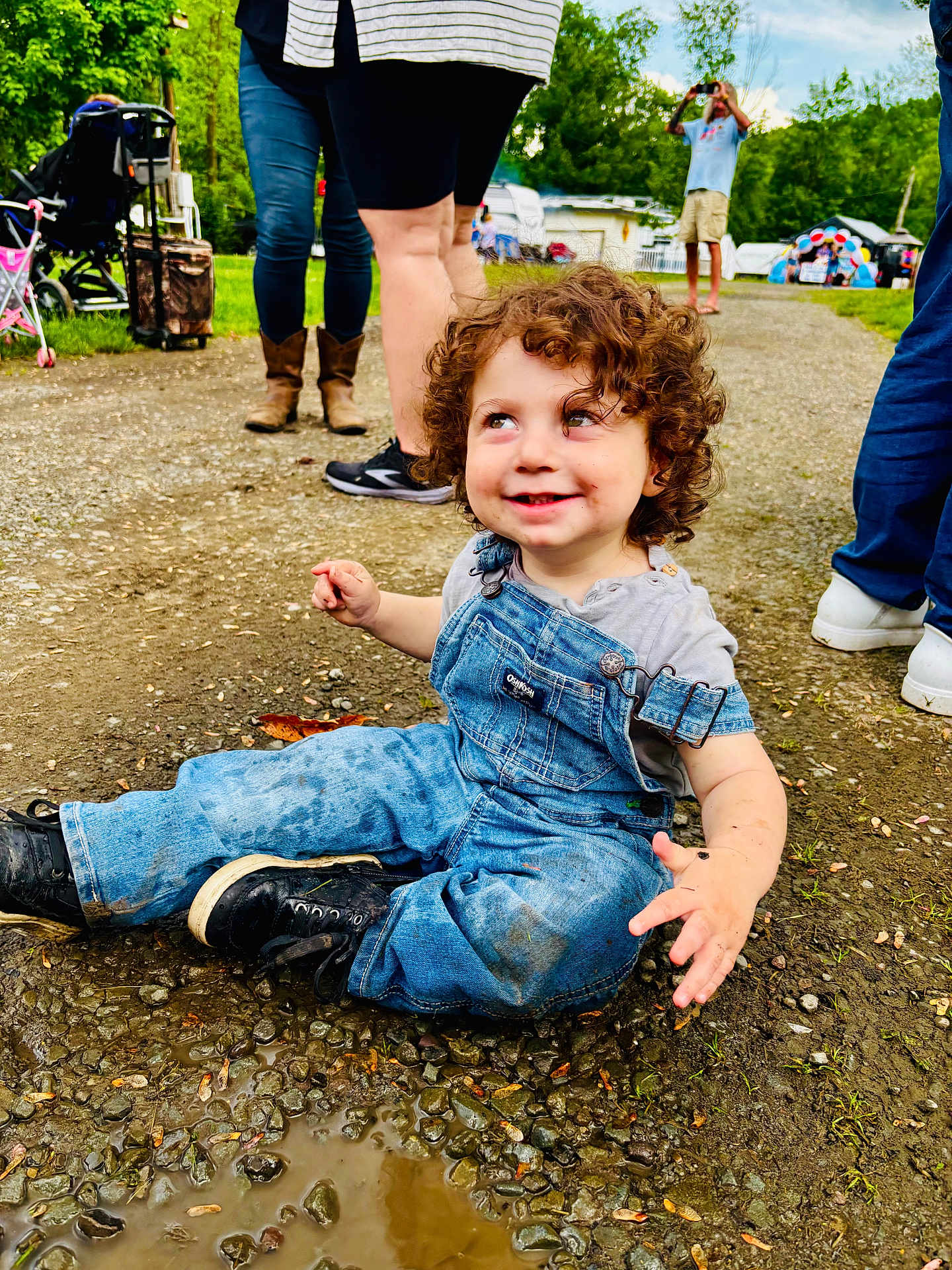 Louie joined the competition — help win amazing prizes! toddler, child, muddy_clothes, denim_overalls, curly_hair, outdoor, gravel_path, puddle, smiling, happy, people, legs, boots, shoes, nature, grass, trees, daylight, casual_clothing, playful
